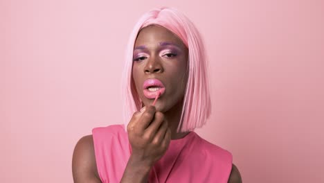 extravagant black transgender applying lip gloss in studio against pink background
