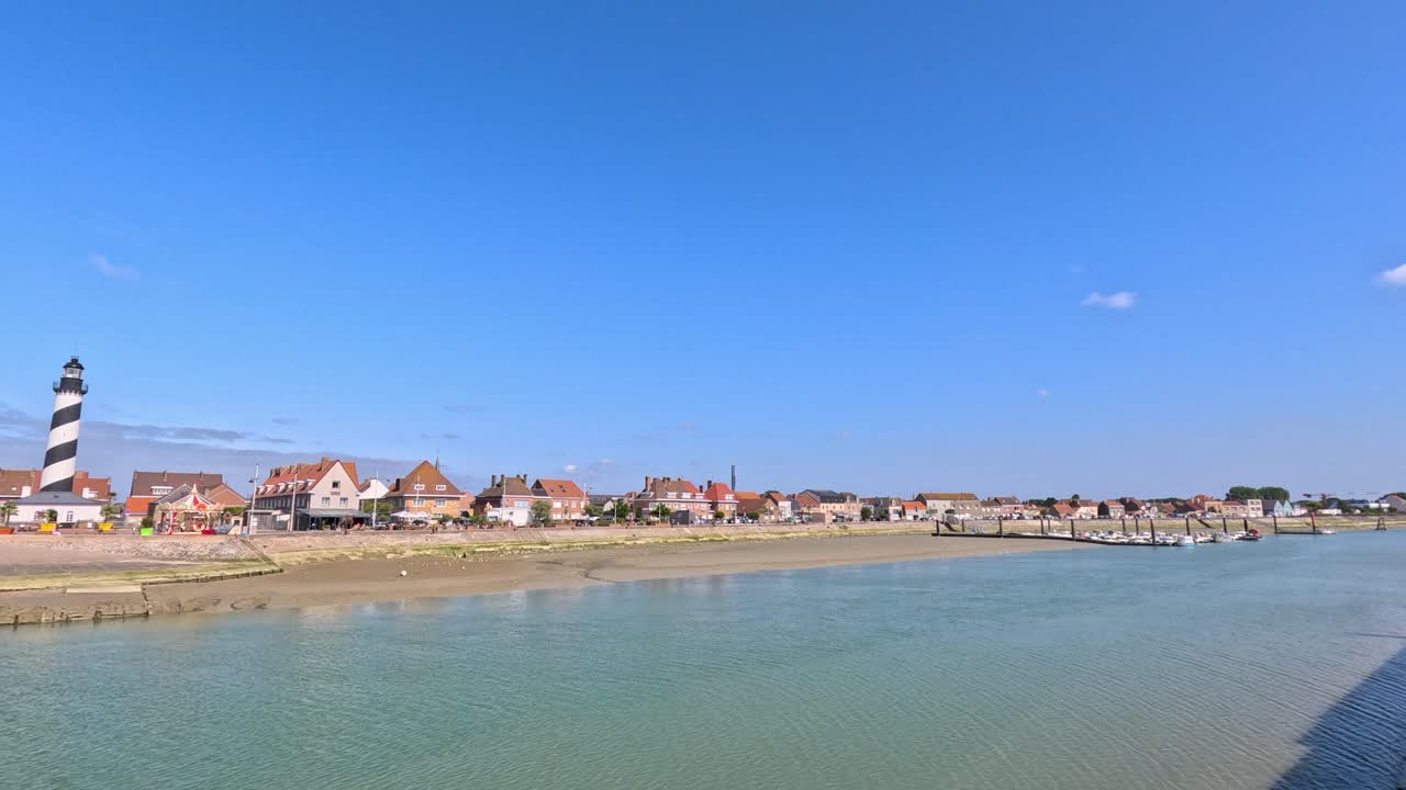 Wide daytime pan of Gravelines coastline with striped lighthouse, waterfront houses, and calm sea