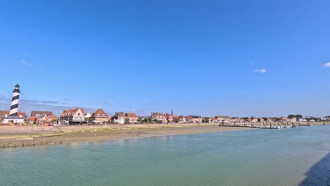 wide daytime pan of gravelines coastline with striped lighthouse, waterfront houses, and calm sea