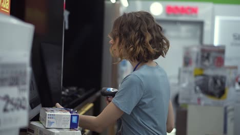 Curly-haired-concentrated-woman-seller-consultant-in-household-department-carry-and-scan-by-barcode-stationery-goods-in-the-boxes,-standing-in-front-the-screen.-Slow-motion.-Side-view