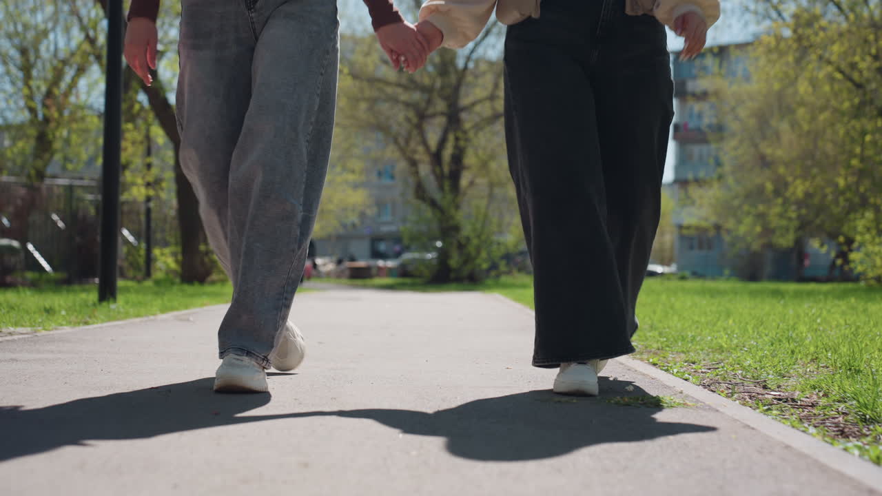 햇빛이 비치는 포장도로를 따라 운동화와 청바지를 입은 백인 여성과 아시아 여성이 걷는 모습 클로즈업, 리드미컬한 발소리, 긴 그림자, 멀리 보이는 보행자와 녹지가 있는 캐주얼한 도시 공원 산책로