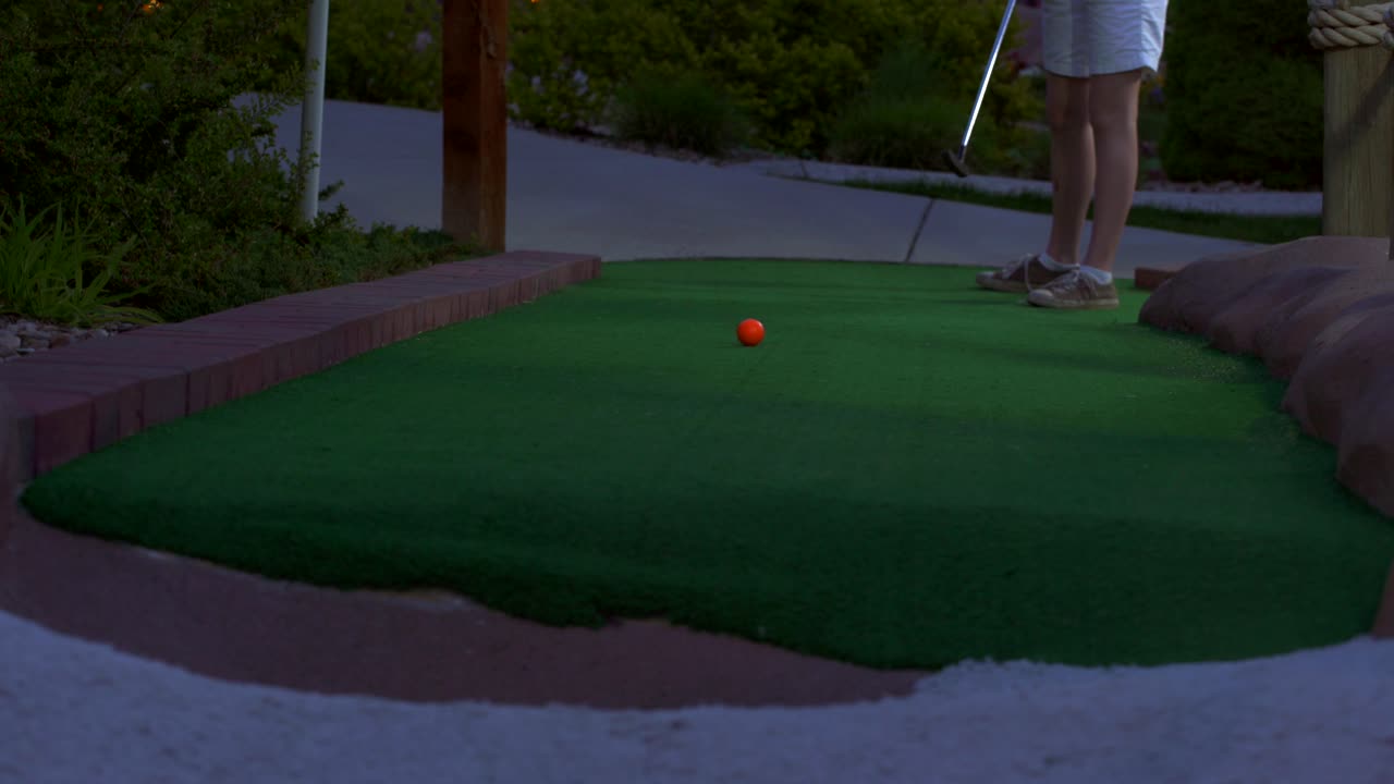 A caucasian girl mini golfing an orange golf ball hitting it towards the camera