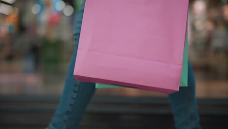 close-up of a pink shopping bag held in someone's hand, with a mint-colored bag slightly visible, set against a vibrant shopping mall background with colorful reflections and bustling activity