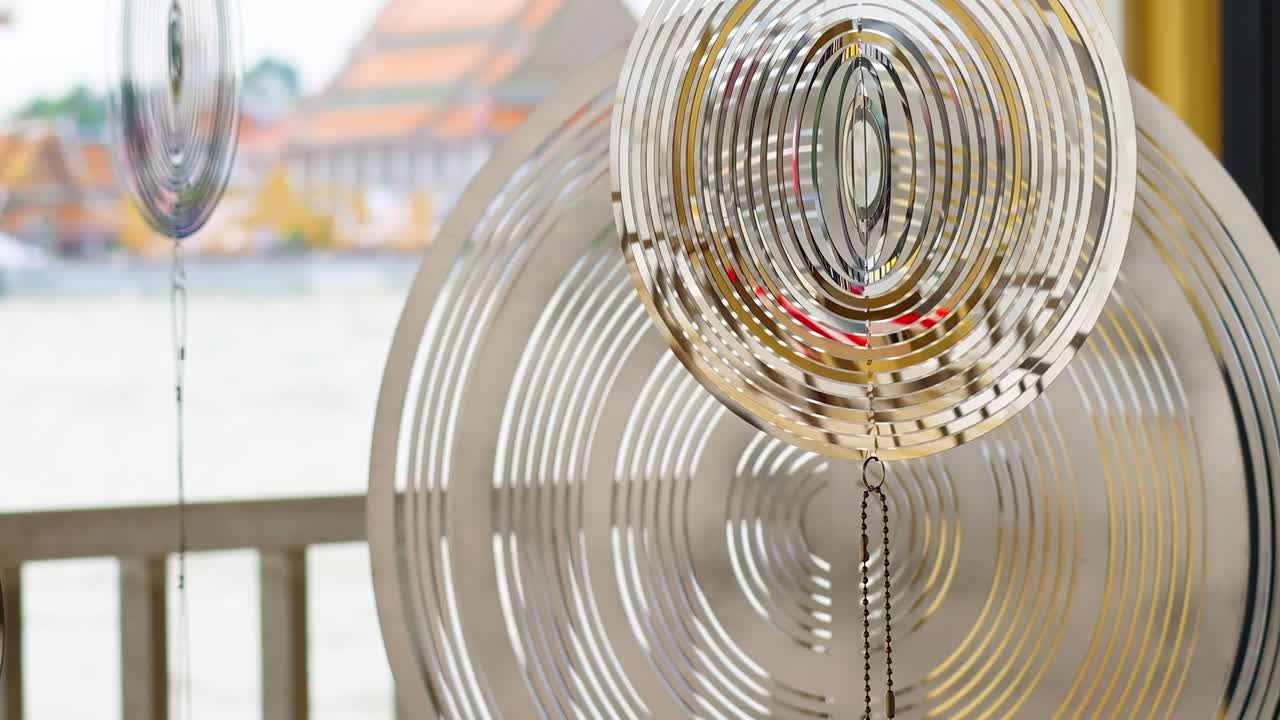 A metal wind chime spins gracefully, reflecting light with a riverside backdrop in the distance.