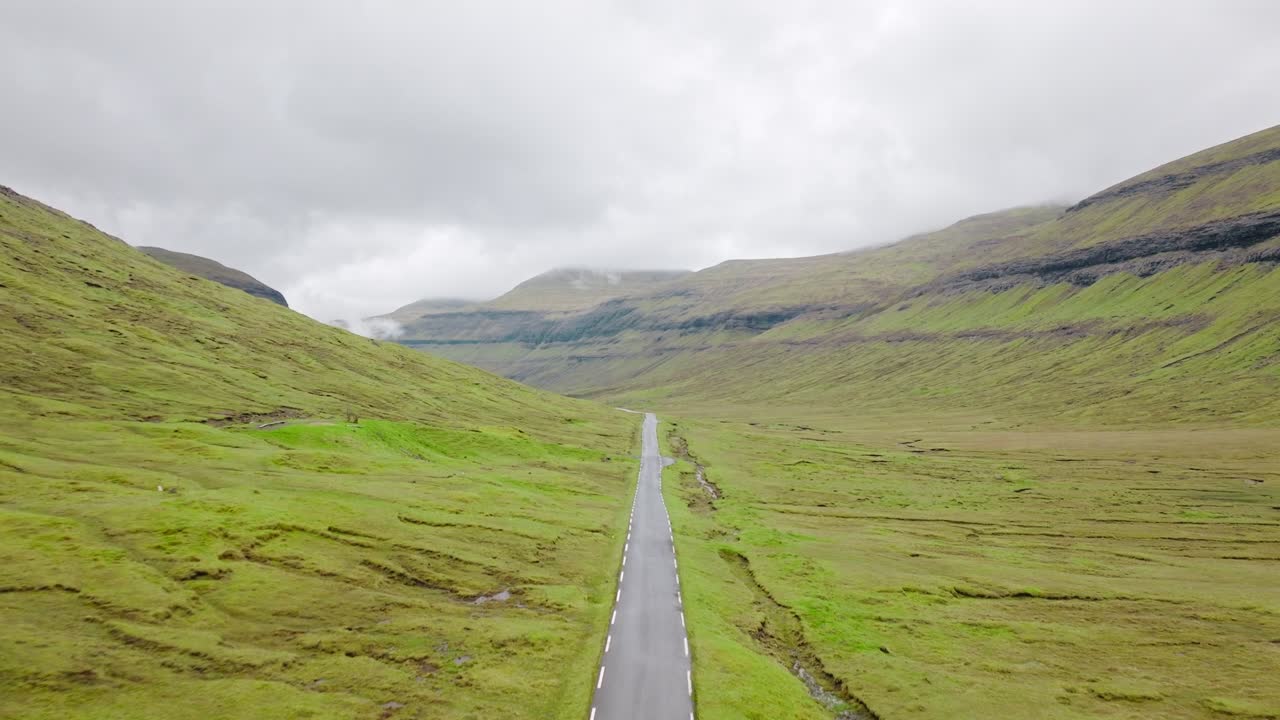 A long winding road cuts through green Faroe valleys, under a cloudy sky, serene