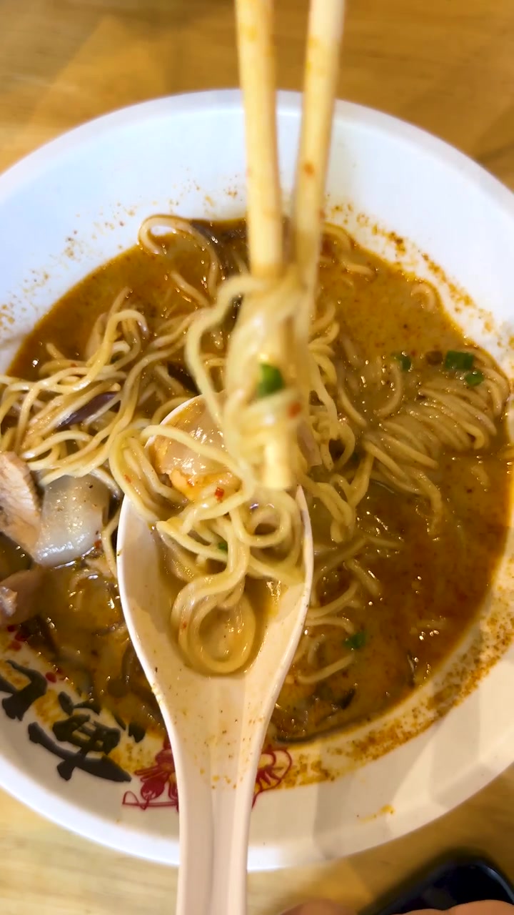 Una mano usa le bacchette per sollevare i noodle ramen da una ciotola in un ambiente di ristorante giapponese ben illuminato.