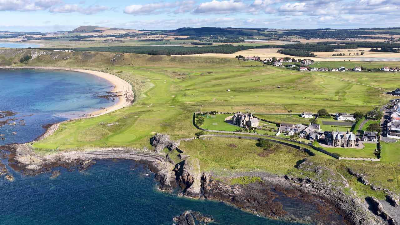 Drone glides above green golf course, rugged cliffs, and coastline under bright daylight in Fife