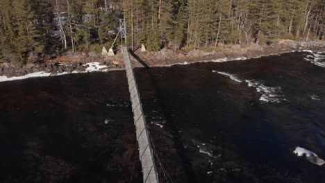 Revele-Una-Toma-Aérea-De-Un-Puente-Colgante-En-Mårdseleforsen,-Cruzando-El-Río-Vindelälven,-Ubicado-En-El-Norte-De-Suecia,-Revelando-Un-Hermoso-Cauce-Con-Altos-Y-Exuberantes-Pinos-Centenarios