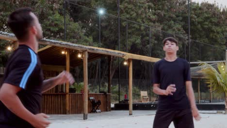 two men playing volleyball on a sandy court at dusk