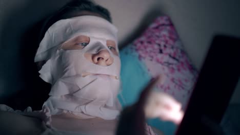 young woman in white sheet mask lies on blue flowered pillow