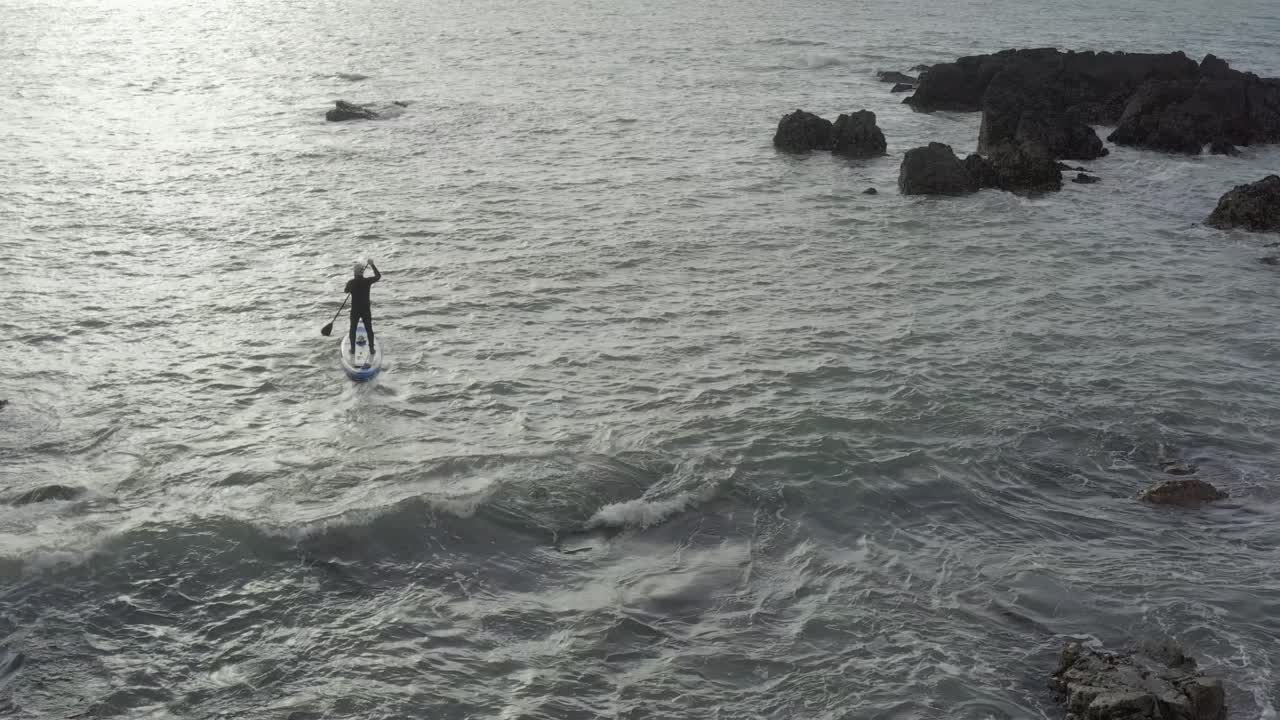 la antena sigue a un pasajero de paddle masculino en un océano agitado cerca de rocas afiladas