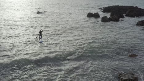 die luftaufnahme folgt einem männlichen paddelboarder auf dem unruhigen meer in der nähe scharfer felsen