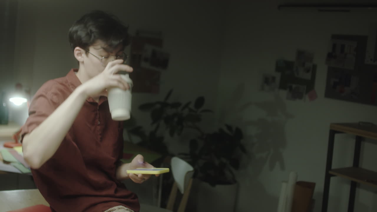 A young person with glasses relaxes or studies in a dimly lit room, holding a cup and looking at a phone
