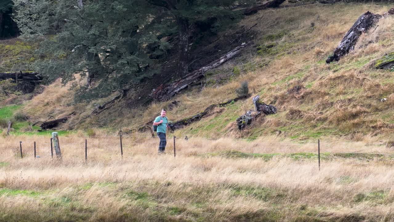 Person with backpack walks beside fence through grassy field, overcast daylight, steady wide shot