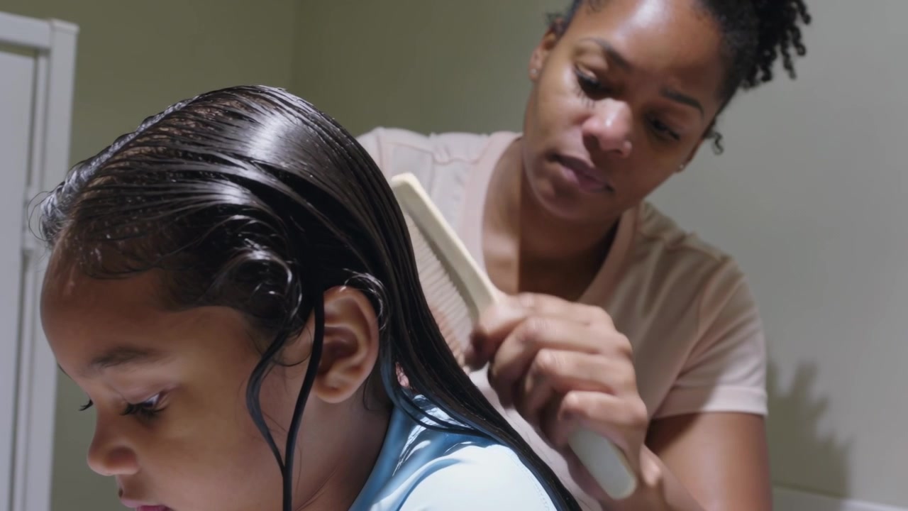 Reaching with wide-tooth comb, mother detangling daughter wearing light blue top's wet hair at home