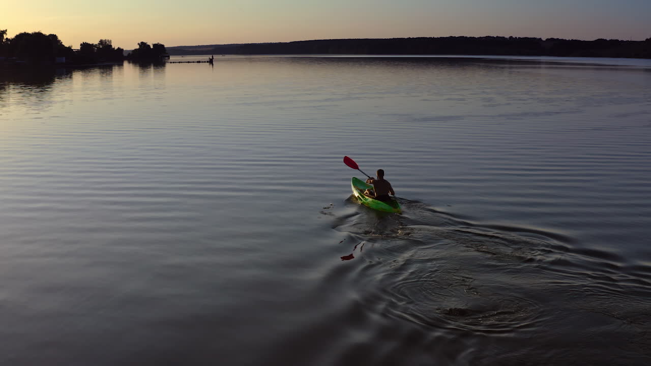 Young man kayaks on lake at sunset. Alone man riding on a rowing boat on the lake at dusk time. Man in a boat with oars in the evening. Aerial view.