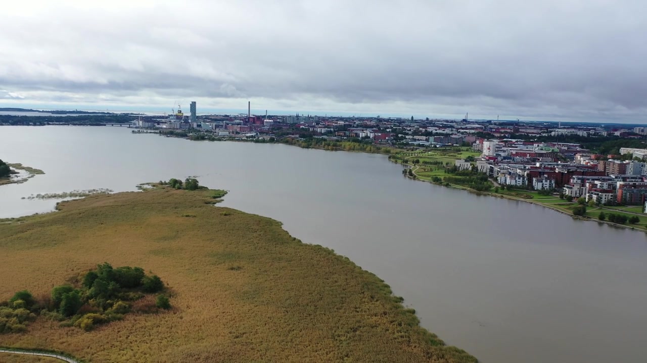 Ampia ripresa aerea dello skyline urbano di Helsinki con i grattacieli di Kalasatama e la centrale elettrica di Hanasaari attraverso la baia di Vanhankaupunginselkä. Paesaggio costiero scenografico sotto un cielo cupo e nuvoloso