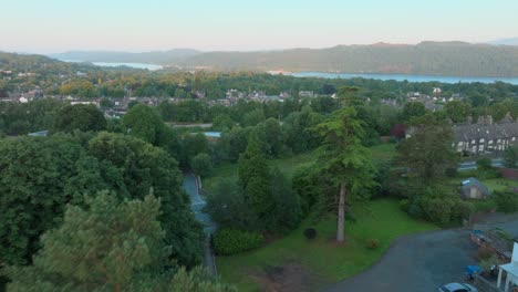 flying through trees revealing rural town next to large lake at sunrise. summer. windermere, cumbria, uk