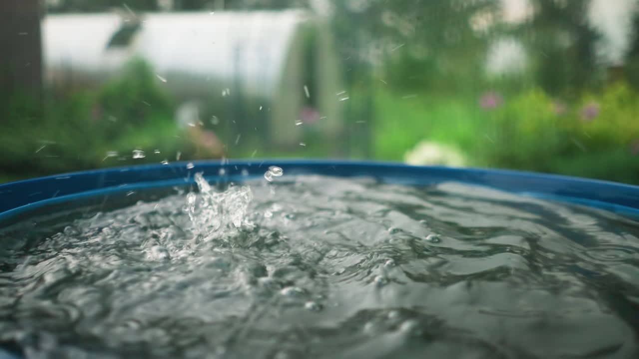 Close-up of water dripping into blue water drum, creating ripples and splashes, background features blurred garden scenery with greenery, flowers, and outdoor elements