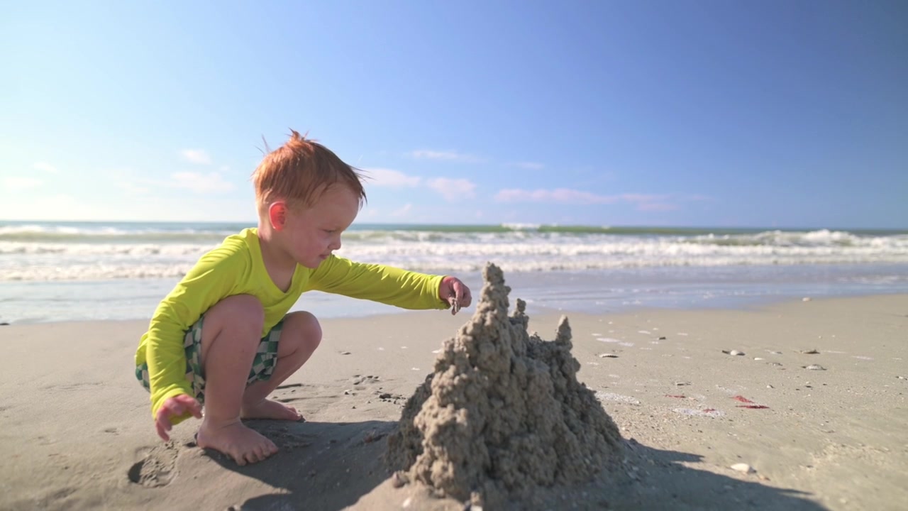 Young boy in bright yellow long-sleeve shirt focuses on crafting a sandcastle on the beach, with the ocean waves rolling in and the sun shining brightly overhead, creating a cheerful atmosphere