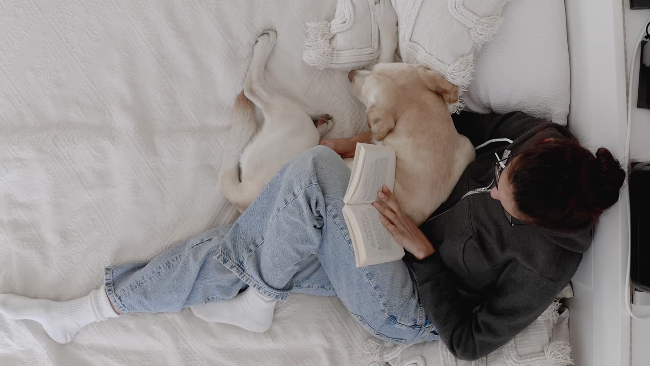 Top-down view of woman sitting on bed reading a book while hugging her dog. Peaceful moment showing human-animal bond, cozy home, love for pets, relaxation, lifestyle, and companionship.
