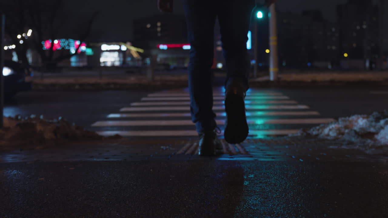 Low angle rear view follow of male in jeans and black boots crossing city street at night