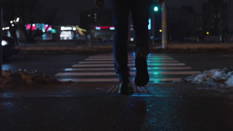 low angle rear view follow of male in jeans and black boots crossing city street at night