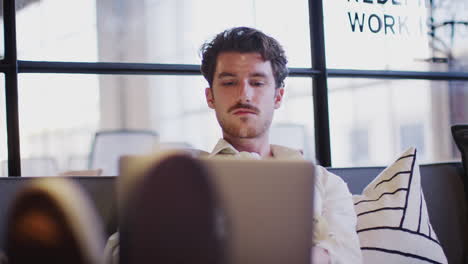 Young-white-man-working-on-a-laptop-sitting-in-an-office-lounge-area,-close-up,-rack-focus