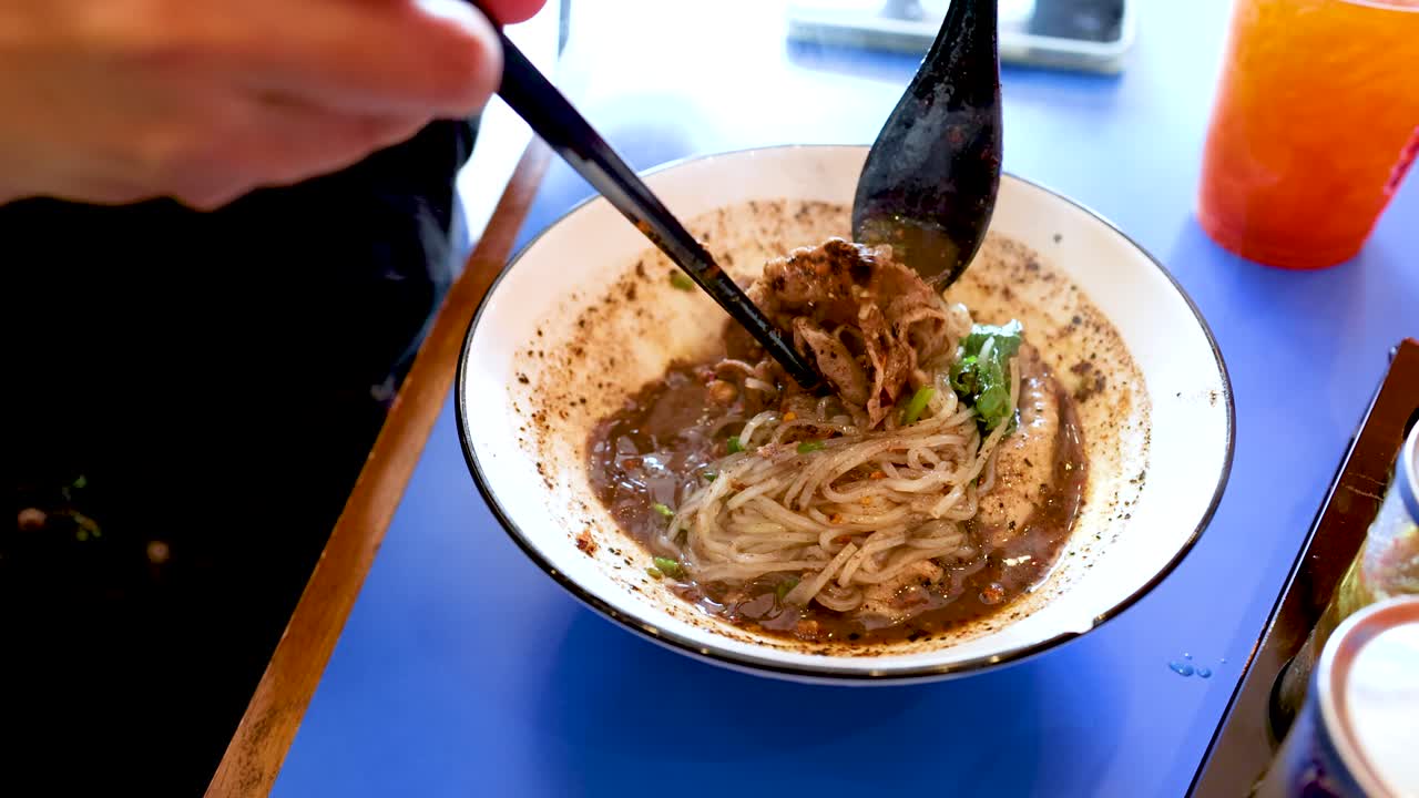 Primo piano di mani che usano bacchette e cucchiaio per mangiare una zuppa di noodle tailandese piccante in stile barca