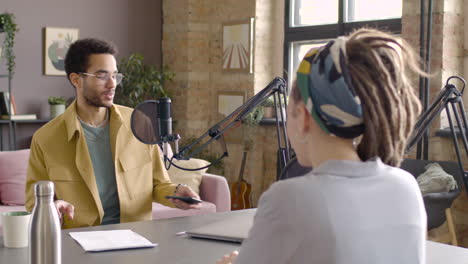 Young-african-american-man-sitting-at-a-table-with-microphones-and-computer-to-record-a-podcast-with-a-caucasian-woman