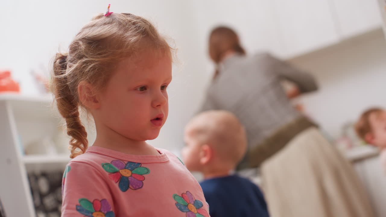 Close up of kindergarten child in pink shirt with flower design standing indoors while teacher in background organizes kitchen items, other children moving around