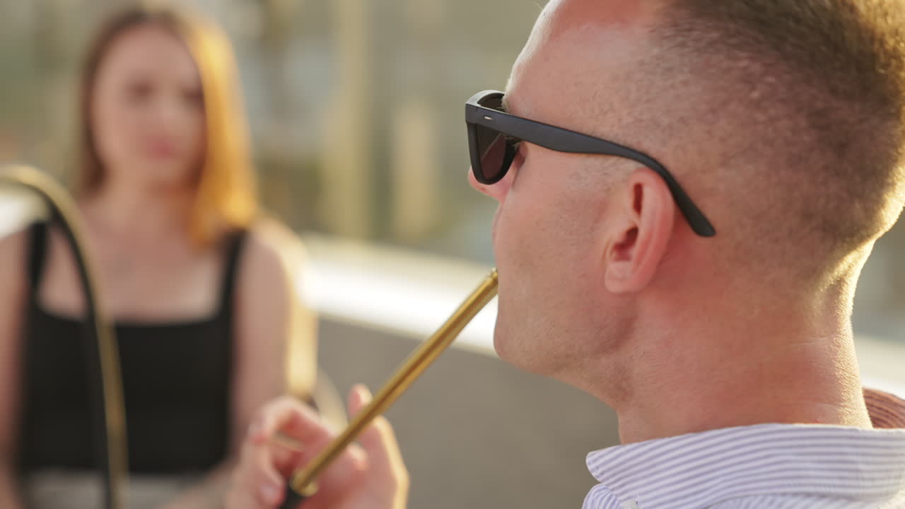 Calm Caucasian man wearing sunglasses smoking hookah sitting in a café outdoors. Girls sitting opposite at backdrop in blur.