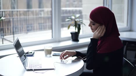 Attraktives-Mädchen-Mit-Traditionellem-Arabischen-Hijab-Arbeitet-An-Etwas-Auf-Ihrem-Laptop-Und-Steckt-Sich-Dann-Kopfhörer-In-Die-Ohren.-Innenaufnahmen-Einer-Wunderschönen-Frau