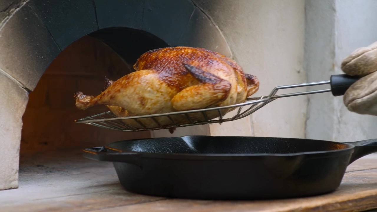 Pulling gloved hand sliding wire rack under roast chicken from masonry oven, draining into skillet
