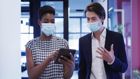 diverse businesswomen wearing face masks talking using digital tablet walking through office