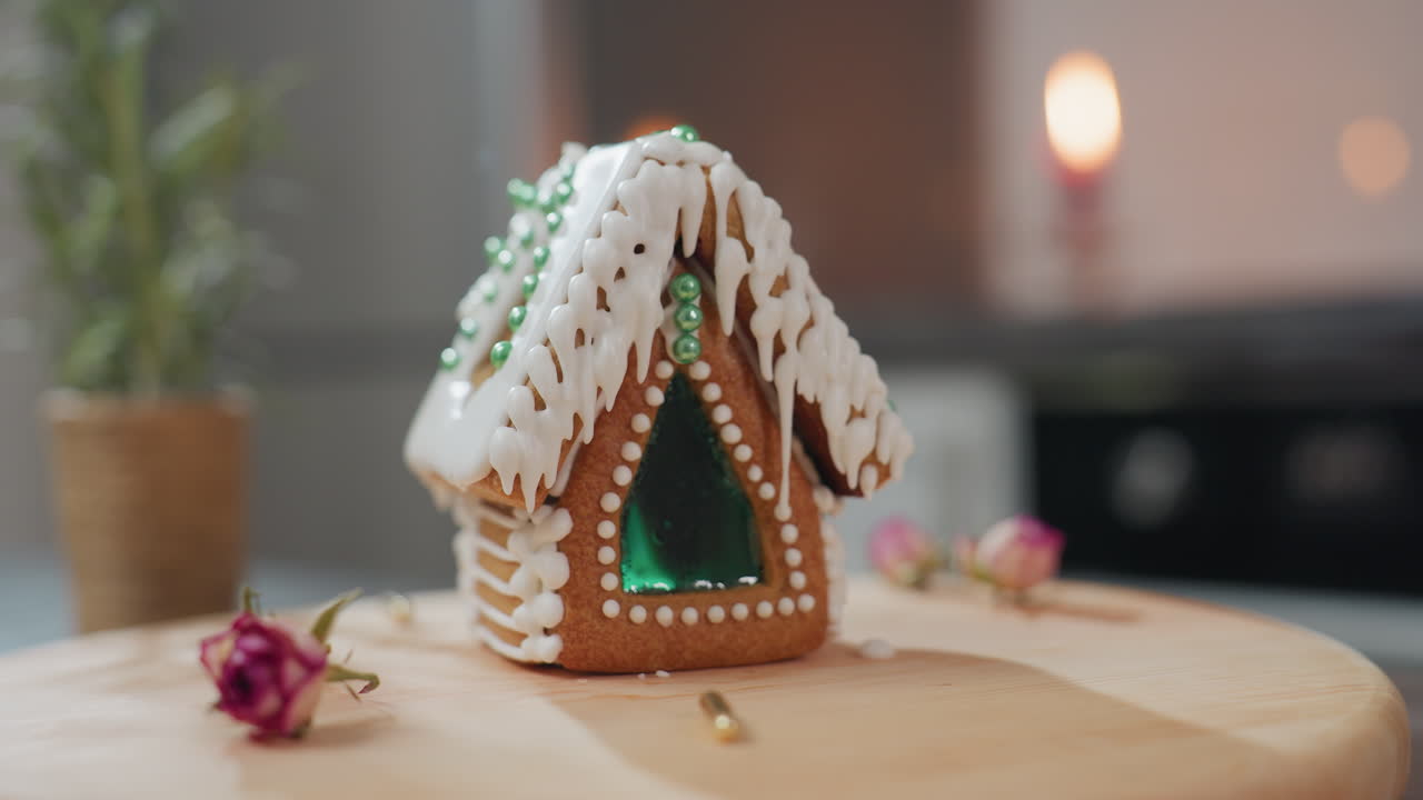 casa de pan de jengibre bellamente decorada con hielo blanco, cuentas verdes, y una ventana translúcida se sienta en una bandeja de madera, galletas, planta en olla, y una vela ardiente