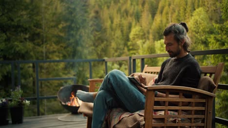 a man with yellow sits on a sofa and works on a tablet on the balcony of a country house overlooking the mountains and coniferous forest. spiritual break and picnic away from the noisy city