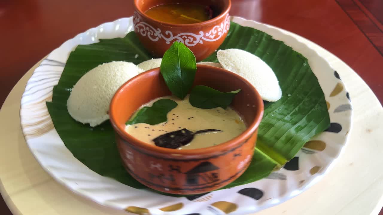 Idly served with sambar and chutney is topped with curryleafs on a rotating table