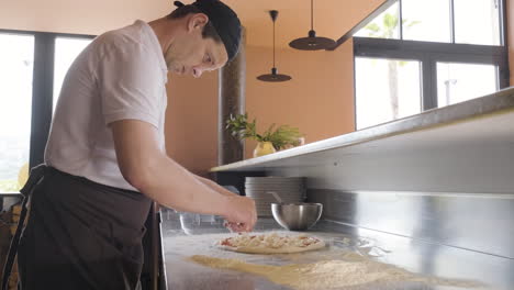 side view of a chef spreading cheese on pizza dough on a restaurant kitchen countertop