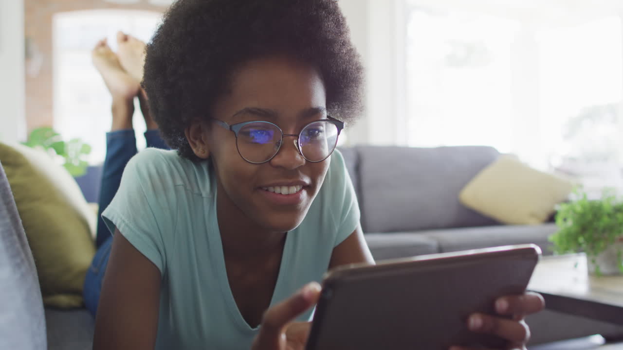 Happy african american teenage girl lying on sofa, using tablet