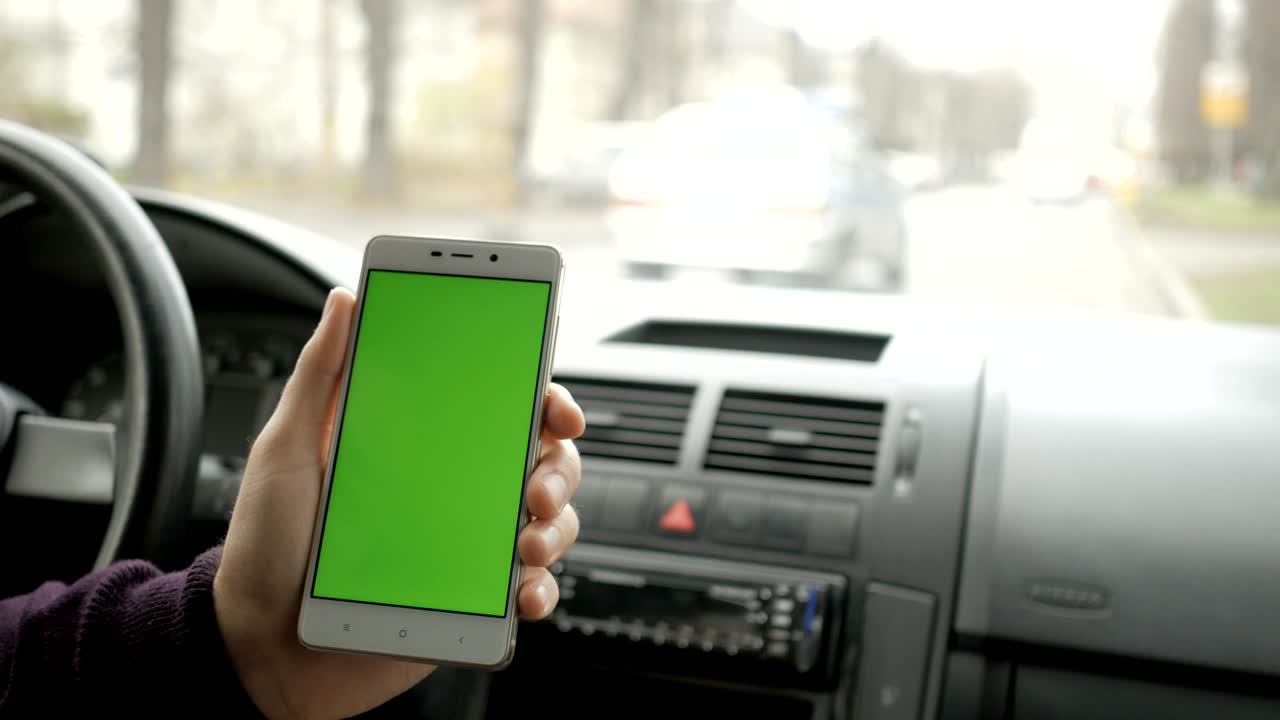 una mano sosteniendo un teléfono inteligente con pantalla verde en blanco en el coche para la dirección, masaje, ubicación, negocio. hombre se sienta en un coche moderno y trabaja en teléfono inteligente - pantalla verde - primer plano. llave de croma.