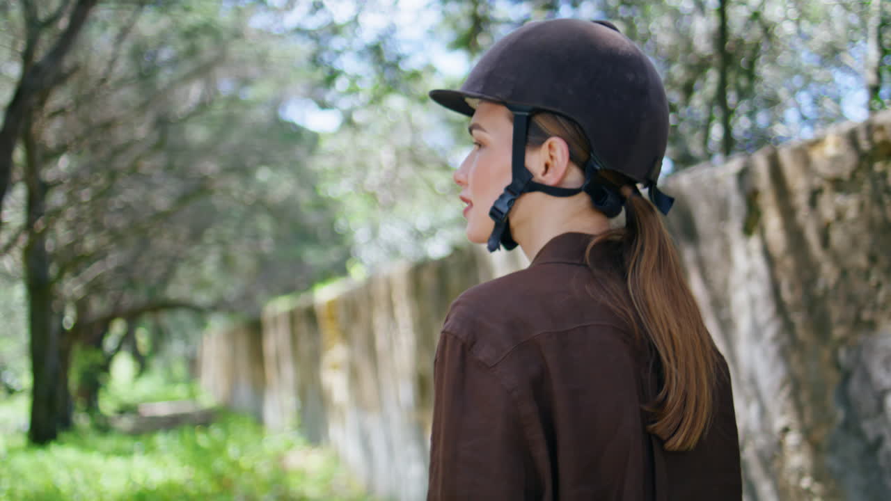 Jockey lady posing stoned wall portrait. Serene cowgirl in helmet walking garden