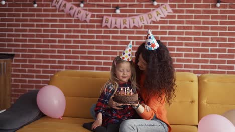 madre sosteniendo en las manos un pequeño pastel de chocolate de cumpleaños, hija soplando las velas