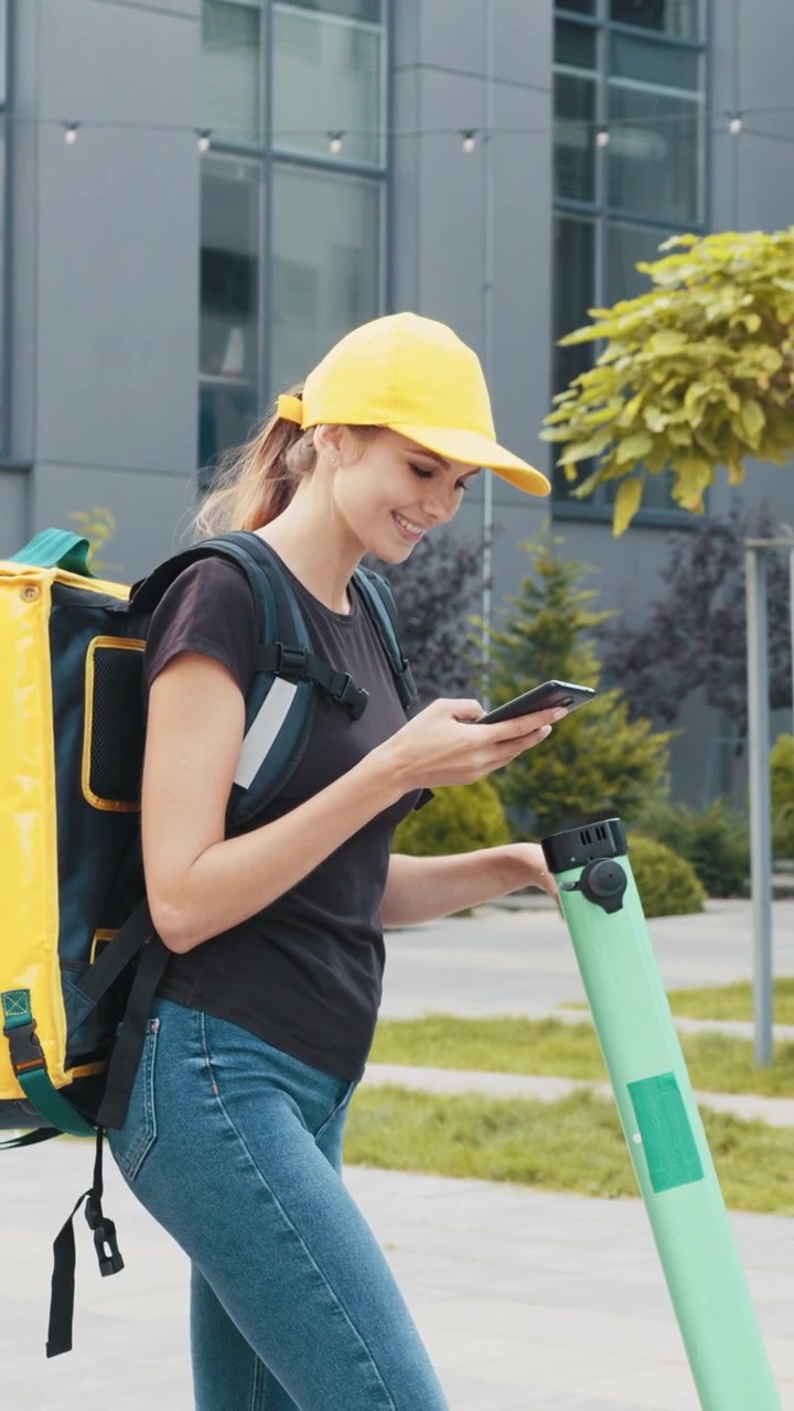 A smiling delivery woman in a yellow cap checks her phone while standing next to her scooter ready