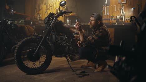 Side-view-of-a-confident-mechanic-in-a-plaid-shirt-inspecting-motorcycle-parts-during-repairs-in-his-workshop-studio