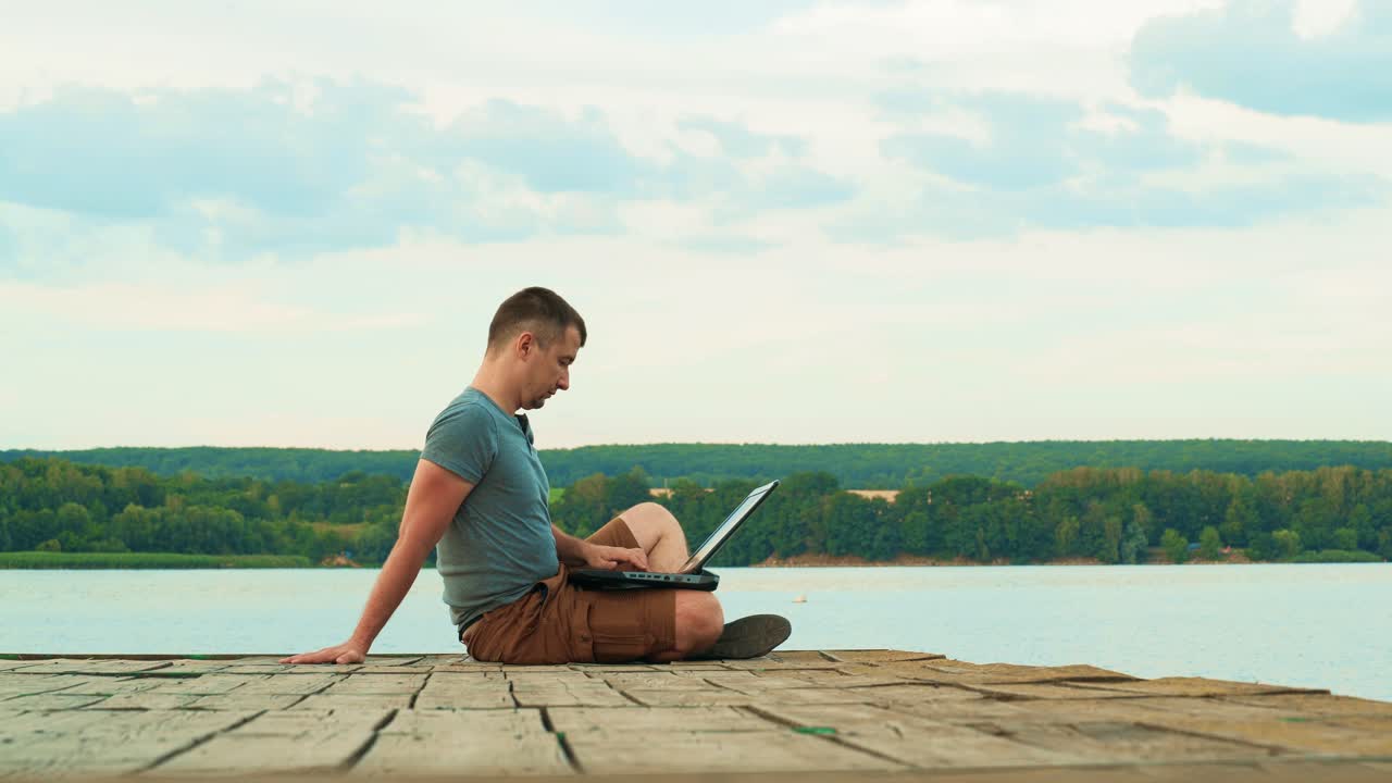Strong man sitting calmly near the river and working on his digital gadget under the blue sky. Beautiful landscape of nature in summer and a man sitting on a wooden bridge with a laptop.