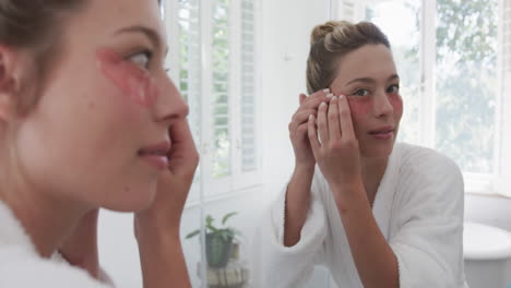 Happy-biracial-woman-applying-under-eye-patches-looking-in-mirror-in-bathroom,-slow-motion