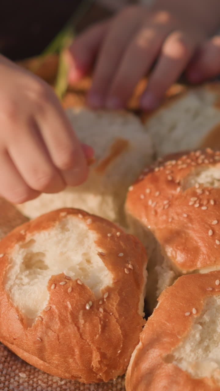 Close-up shot of children's hands pinching and eating freshly baked sesame bread rolls on a checkered picnic blanket. The scene captures a simple and peaceful outdoor moment