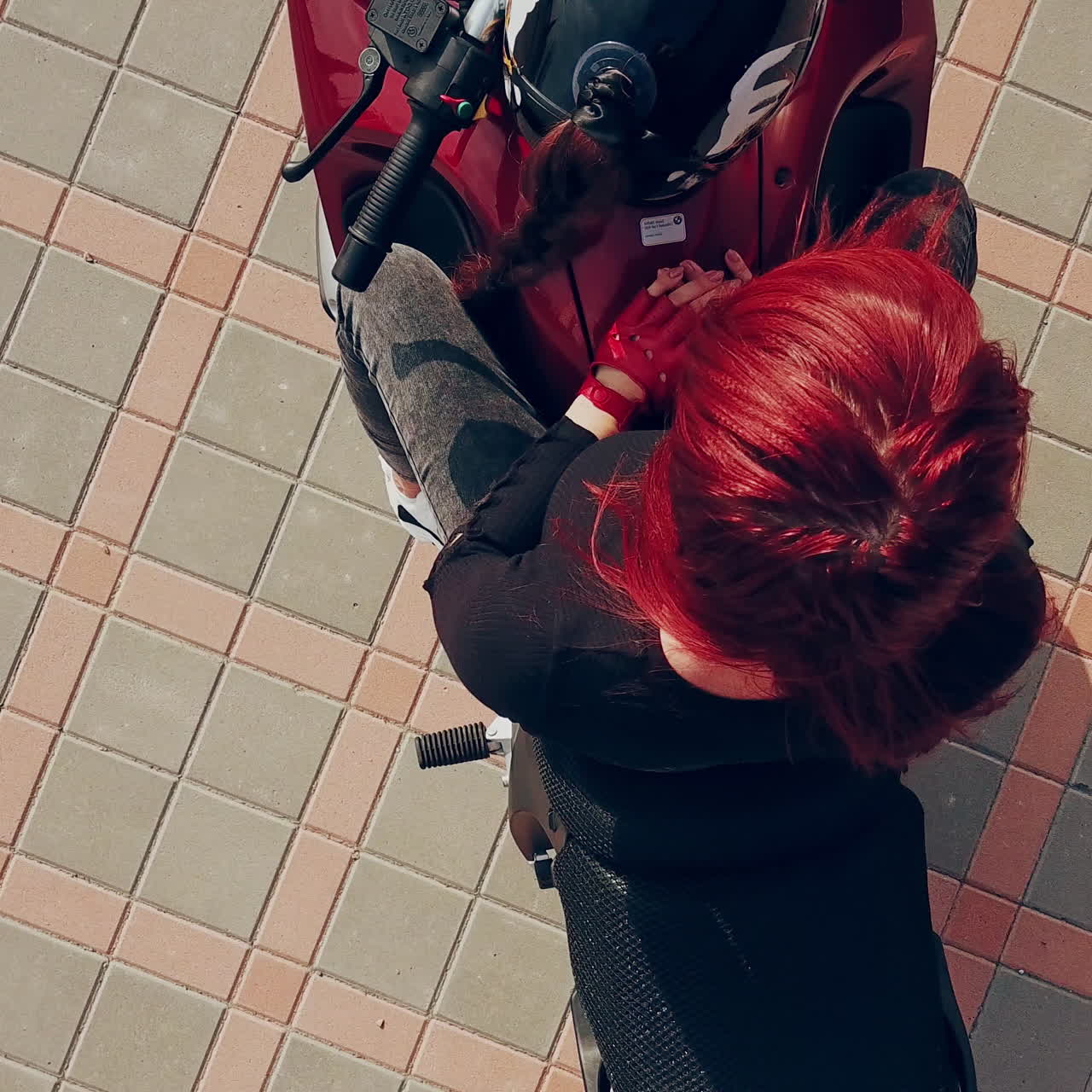 Red-haired young woman sitting on a red motorcycle and lying on it over the background of paving tile. Camera moves up.