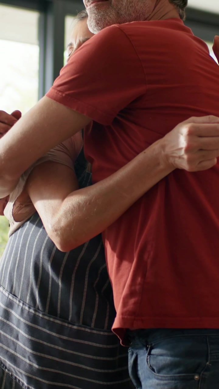 Video verticale: Uomo che si avvicina a una cuoca casalinga con grembiule a righe, che abbraccia e tiene un cucchiaio vicino alla finestra.